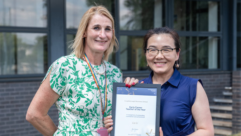 Two people standing in front of a building with large windows and steps; one holds a framed 